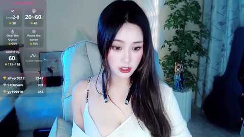 Media: Video of a young Asian woman with long black hair, wearing a white off-shoulder top, seated in a modern, dimly lit room with a plant and white curtains.