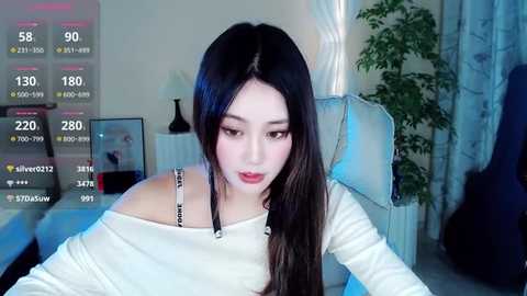 Media: Video of a young East Asian woman with long black hair, wearing a white off-shoulder top, seated in a dimly lit room with a guitar, plants, and a white chair.