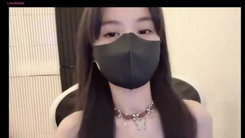 Media: Video of an East Asian woman with long, straight black hair, wearing a black face mask, silver necklace, and black top, indoors with beige walls and a black screen in the background.