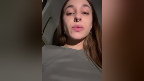 Media: Video of a young woman with fair skin, brown hair, and a nose piercing, wearing a gray top, taken indoors with soft lighting.