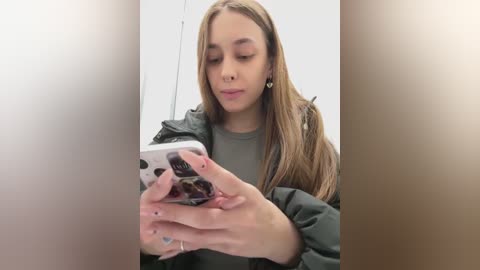 Media: Video of a young, light-skinned woman with long, straight brown hair, wearing a grey hoodie, taking a selfie with a white phone. Background is blurred, showing a plain white wall.