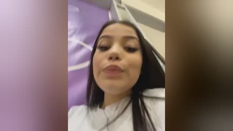 Media: Video of a young woman with medium skin tone, long dark hair, and full lips making a pouty face, wearing a white top, in a brightly lit, modern interior with purple accents.