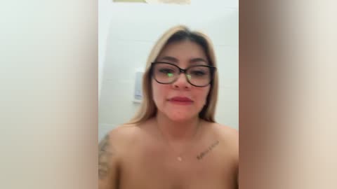 Media: Video of a topless woman with long blonde hair, wearing black-rimmed glasses and a tattoo on her left arm, standing in a bathroom with white tiles.