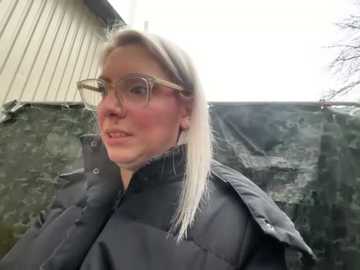 Media: Video of a middle-aged woman with long blonde hair, wearing glasses and a dark puffy coat, standing in front of a gray, textured wall and a partially visible building.