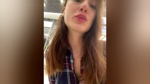 Media: Video of a young Caucasian woman with long brown hair, wearing a red and blue plaid shirt, puckering her lips for a kiss. Background shows a tiled ceiling and blurred indistinct objects.