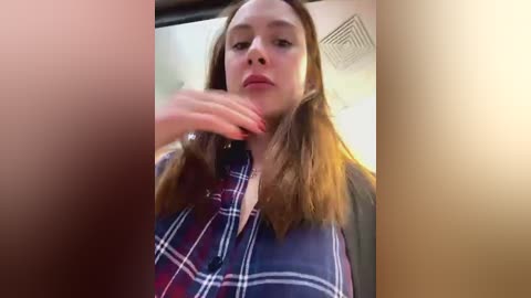 Media: A video of a young woman with fair skin, long brown hair, and red nail polish, wearing a blue plaid shirt. She is inside a car, with a blurred background showing a vent.