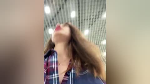 Media: Video of a woman with long brown hair, wearing a plaid shirt, tilted head back, with a blurred, modern, industrial ceiling in the background.