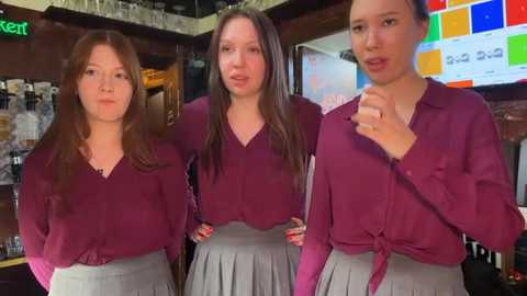 Media: Video of three young women with light skin, wearing maroon tops and gray skirts, standing in a bar. Background includes a colorful menu board, bar shelves, and glasses.