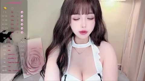 A video of a young, fair-skinned woman with long, wavy brown hair and blunt bangs, wearing a white lingerie top with black straps, posing indoors with soft, pastel-colored background.