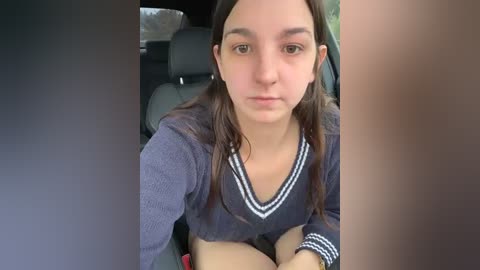 Media: Video of a young woman with long dark hair, wearing a navy blue sweater, sitting in a car with black seats, looking serious.