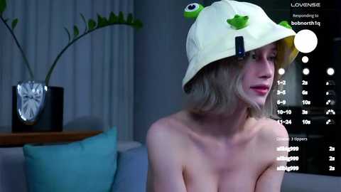 Media: Video of a topless, blonde woman with fair skin, wearing a green frog hat, sitting on a couch. Background features a black clock, white curtains, and a green plant.