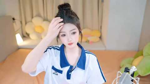 Media: Video of a young Asian woman with fair skin and long dark hair in a bun, wearing a white polo shirt with blue stripes and sleeves. She's touching her head, looking concerned. Background features a bed with green and yellow stuffed animals, beige curtains, and a white lamp.