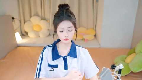 Media: Video of a young East Asian woman with dark hair in a bun, wearing a white polo shirt with blue trim, seated at a desk in a cozy, softly lit room with floral decorations and plush toys.