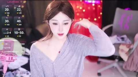 Media: Video of a young Asian woman with light skin, long brown hair, and fair complexion, wearing a light gray cardigan, adjusting her hair in a dimly lit room with a red light in the background.