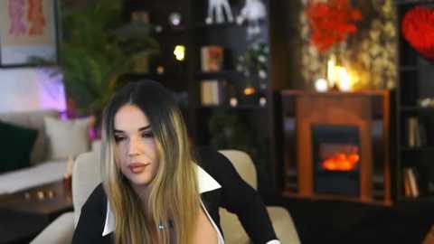 Media: Video of a fair-skinned woman with long, blonde-streaked black hair, wearing a black jacket with white collar, sitting in a modern living room with dark furniture, fireplace, plants, and abstract art.