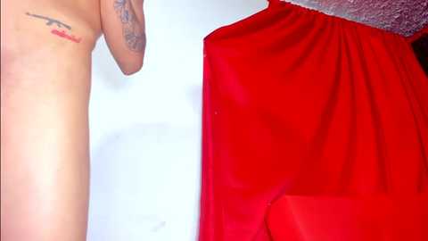 Media: Video of a topless person with a small tattoo on the right shoulder, standing beside a vibrant red curtain with a white wall in the background.