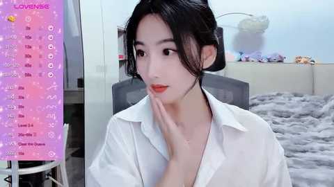 Media: Video of an East Asian woman with fair skin, black hair, wearing a white shirt, looking at her hand in a cozy bedroom with a gray blanket and stuffed animals.