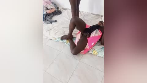 Media: Video of a naked black man, lying on a tiled floor, wearing a pink and black cloth, surrounded by scattered clothes and a white sheet in a dimly lit room.