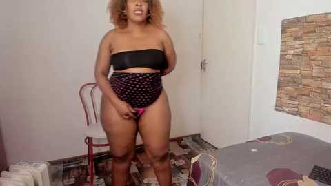 Media: Video of a plus-size Black woman with curly hair, wearing a black strapless top and high-waisted black panties with pink polka dots, standing in a sparsely decorated bedroom with a wooden wall, red chair, and bed.