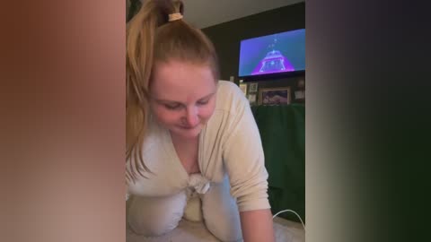 Media: Video of a smiling, fair-skinned woman with long blonde hair tied in a high ponytail, wearing a light beige cardigan, leaning forward on a bed in a dimly lit room.