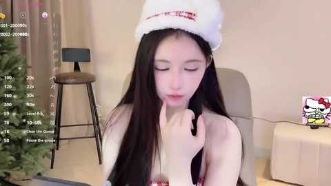 Media: Video of an Asian woman with long black hair wearing a Santa hat, seated in a beige chair, holding a black pen to her lips, in a modern room with a small Christmas tree.