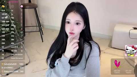 Media: A video of a young Asian woman with long black hair, wearing a gray sweater, sitting on a beige carpet in a room with a white chair and a Christmas tree.