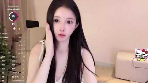 Media: Video of a young East Asian woman with long black hair, wearing a white tank top, sitting on a beige sofa, in a cozy living room with a Christmas tree.