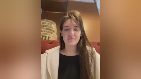 Media: Video of a young woman with long brown hair, wearing a white jacket over a black shirt, sitting on a red sofa in a warmly lit room with a large, decorative, beige-and-black scroll patterned cushion in the background.