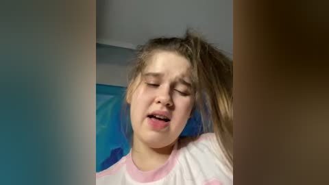 Media: Video of a young Caucasian girl with fair skin and long, messy brown hair, wearing a white shirt, lying down with eyes closed, looking relaxed in a room with blue and white walls.