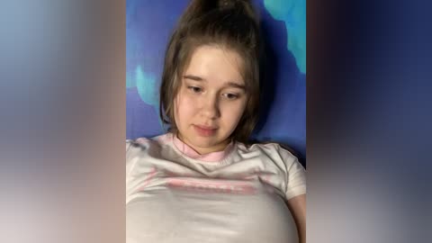 Media: A video of a young Caucasian girl with light skin, brown hair tied in a high ponytail, wearing a white T-shirt with pink text, lying on a blue and purple patterned background.