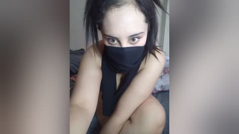 Media: A video of a young woman with light skin and dark hair, wearing a black mask, sitting on a bed with grey sheets. She has a slender physique and a neutral facial expression.