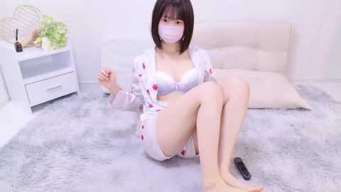 Media: Video of an Asian woman with short black hair, fair skin, and a small frame, wearing a white nurse outfit and face mask, sitting on a fluffy gray rug in a minimalist room with white furniture.