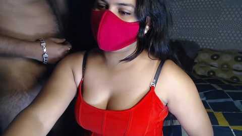 Media: A video of a dark-skinned woman with long black hair, wearing a red face mask and a red velvet dress, sitting on a bed with a patterned blanket.