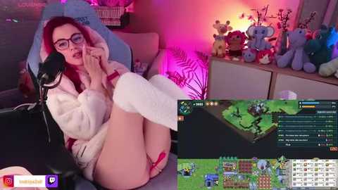 Media: Video of a fair-skinned woman with red hair, wearing glasses, and a white fluffy robe, sitting on a gaming chair, surrounded by plush toys and colorful lights, streaming Minecraft gameplay.