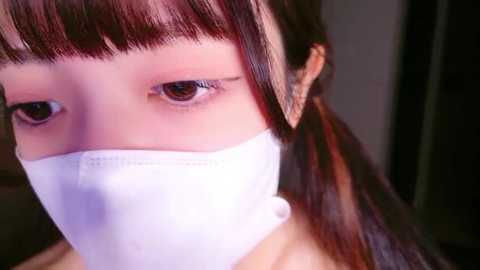 Media: Close-up video of a young woman with light skin, brown eyes, and straight, dark brown hair with bangs. She wears a white face mask, revealing a small portion of her forehead and nose. The background is dimly lit, making the subject stand out.