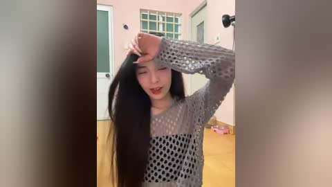 Media: Video of a young Asian woman with long black hair, wearing a grey knit sweater, rubbing her forehead in a hallway with wooden floors and light pink walls.