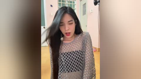 Media: Video of an Asian woman with long black hair, wearing a gray, sheer, textured top, smiling with closed eyes, in a pastel-colored room with wooden flooring and a white door.