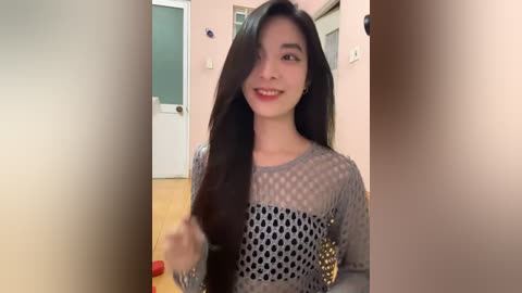 Media: A video of a young Asian woman with long, straight black hair, smiling, wearing a gray mesh top, standing indoors in a dimly lit room.