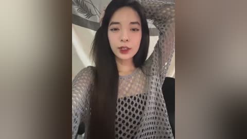 Media: Video of an East Asian woman with long black hair, fair skin, and a slender build, wearing a gray, fishnet sweater. She poses confidently indoors, with a white and black floral-patterned backdrop.