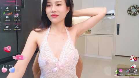 Media: A video of a slender East Asian woman with long black hair, wearing a lacy, pink halterneck top, posing in a modern, well-lit kitchen with white cabinets and a refrigerator.