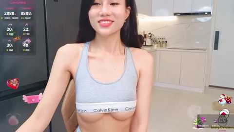 Media: Video of an Asian woman with long black hair, wearing a light grey Calvin Klein sports bra, standing in a modern kitchen.