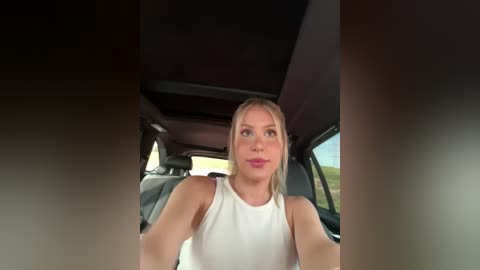 Media: Video of a blonde woman in a white sleeveless top driving a car, with blurred road and greenery visible outside.