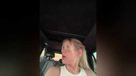 Media: Video of a blonde woman in a white tank top, looking surprised while driving a car. The interior shows dark upholstery and a rear-view mirror.