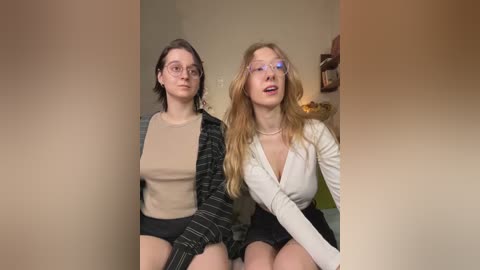 Media: Video of two young women with fair skin, one in glasses, wearing a beige sweater and black shorts, and the other with long blonde hair in a white top, sitting closely indoors.