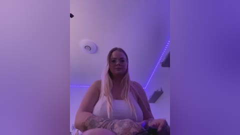 Media: A video of a blonde woman with long dreadlocks, wearing a white tank top, standing in a dimly lit room with purple LED lights and a smoke detector on the wall.