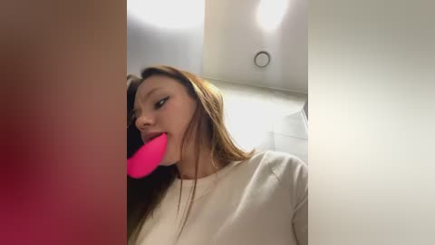 Media: Video of a young woman with straight brown hair, wearing a beige sweater, licking a pink dildo in a dimly lit room with white walls and a recessed ceiling light.