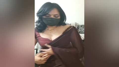 Media: Video of a woman with medium skin tone, wearing a burgundy saree and black mask, adjusting her top, in a dimly lit room with blurred background.