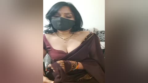 Media: Video of a South Asian woman with medium skin tone, wearing a black mask, dark purple sari with off-shoulder, and multiple bracelets. She holds a small object, likely a phone, in her gloved hand. Background is blurred.