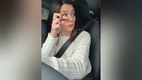 Video of a young woman with long brown hair, wearing a white cable-knit sweater, adjusting her glasses while seated in a car, with a black seatbelt across her torso.