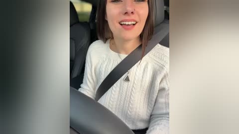 Media: Video of a smiling young woman with light skin and shoulder-length brown hair, wearing a white knitted sweater, seated in a car with a seatbelt across her chest.